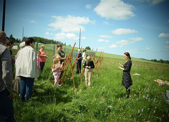 Uczestnicy, stojąc przy sztalugach wysłuchują instruktora, który udziela wskazówek